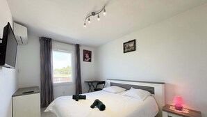 A hotel room with a bed, bedside tables, a TV, a window with curtains, and a ceiling light fixture. - Agde’Loc – The Sunny Cocoon (Vias)