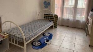 A small, tidy room with a single bed, a small table with books, a fan, and a window with curtains. - Downtown Apartment – 100 m from the Beach (Vendres)