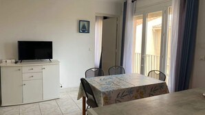 A room with a dining table, chairs, a TV on a cabinet, and a door leading to a balcony. - Downtown Apartment – 100 m from the Beach (Vendres)