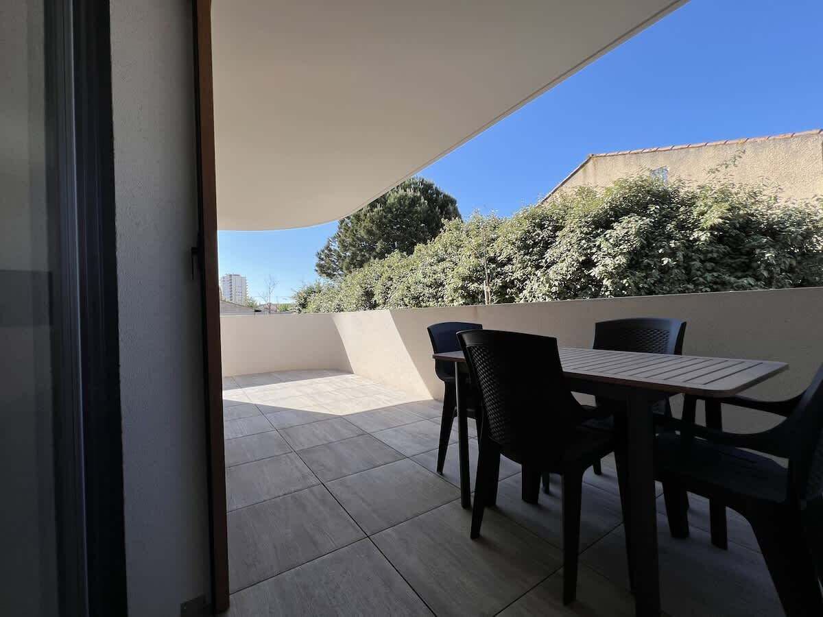 Un espace extérieur aménagé en terrasse, avec une table et des chaises, un coin salon abrité et une vue sur les arbres et les bâtiments en arrière-plan.