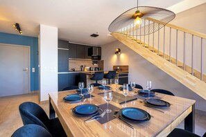 Uma sala de jantar com uma mesa de madeira posta para seis, uma escada e uma cozinha ao fundo.