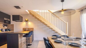A modern kitchen with a central island, a dining area with a wooden table and black chairs, and a staircase with a wooden railing. - Casa Naia - 4-Star Prestige Duplex Valras-Plage (Sérignan)
