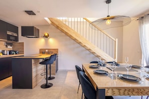 Cozinha moderna com ilha central, área de jantar com mesa de madeira e cadeiras pretas, e escada com corrimão de madeira.