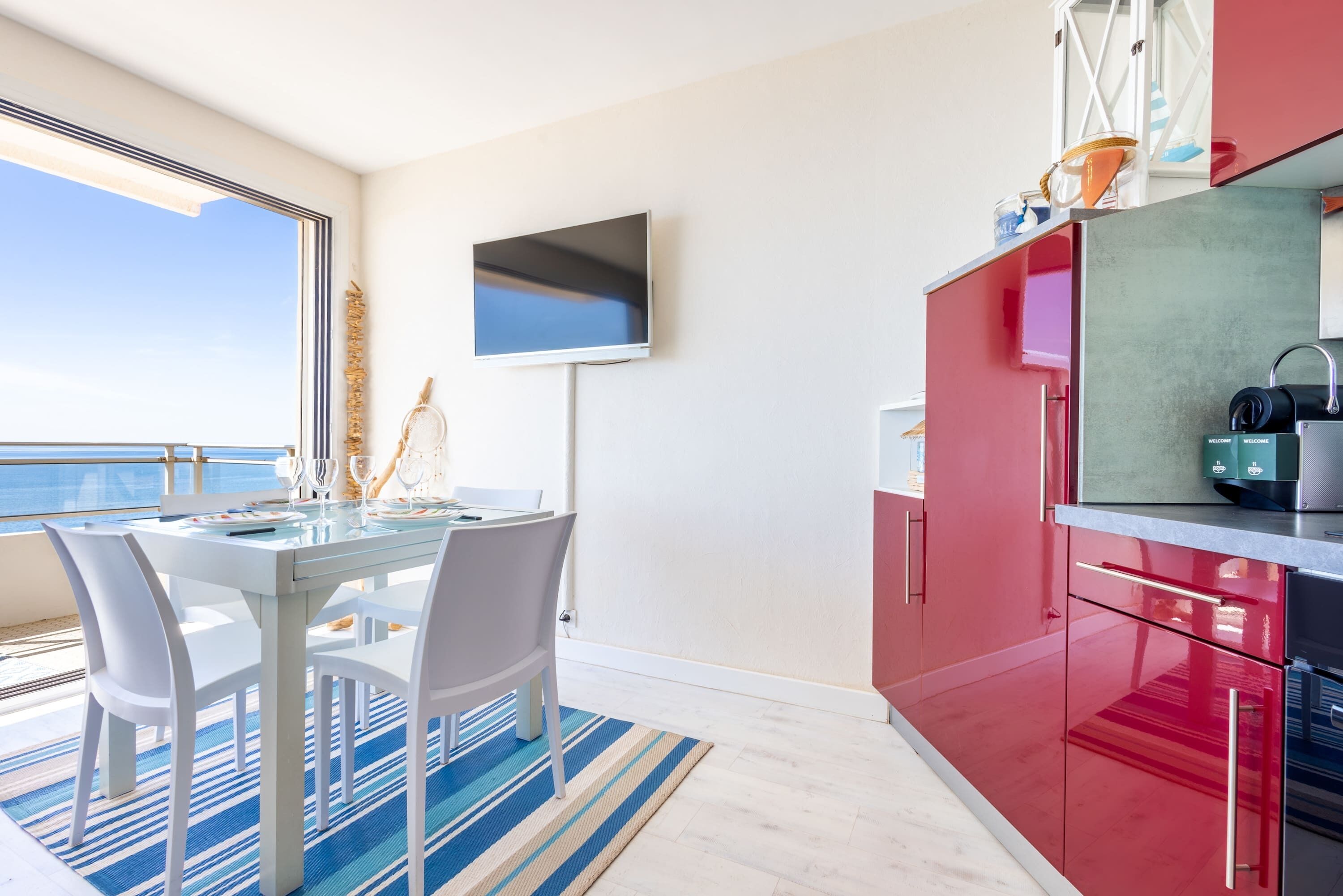 Une cuisine moderne avec des meubles rouges, un téléviseur à écran plat et un coin repas comprenant des chaises blanches et une table en verre.