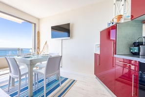 A modern kitchen with red cabinets, a flat-screen TV, and a dining area with white chairs and a glass table. - Bel Horizon – Magnificent apartment with panoramic sea view (Valras-Plage)