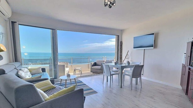 A modern living room with a large window overlooking the sea, a dining area with a table and chairs, and a flat-screen TV mounted on the wall. - Bel Horizon – Magnificent apartment with panoramic sea view (Valras-Plage)