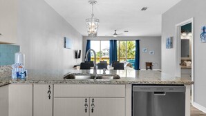 A modern kitchen with granite countertops, stainless steel appliances, and a dishwasher. There is a dining area with a table and chairs visible through the window. - Coastal Cottage (Navarre)