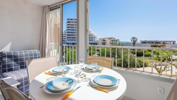 A balcony with a dining table set for two, overlooking a cityscape and a casino. - Valras Apartment with Sea View (Valras-Plage)
