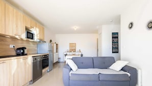 A modern kitchen with wooden cabinets, a microwave, and a dishwasher. There is a grey sofa with cushions in the living area.