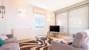 Un salon comprenant un canapé, une table basse, un téléviseur à écran plat, un attrape-rêves fixé au mur et une fenêtre avec des stores.