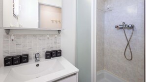 Un baño con un lavamanos blanco, un espejo, una ducha con cabezal de ducha de mano y cuatro cajas de bienvenida.