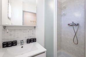 Une salle de bain avec un lavabo blanc, un miroir, une douche avec un pommeau de douche amovible et quatre boîtes d’accueil.