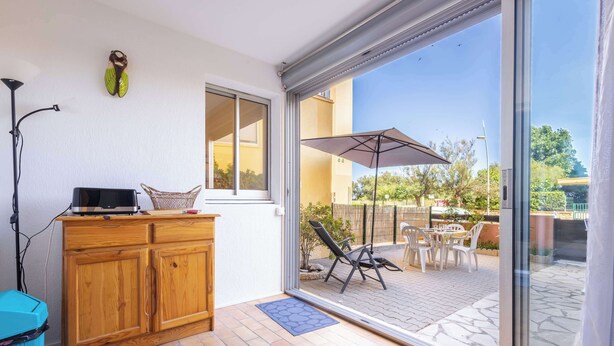 Una habitación con un armario de madera, un televisor, una cesta de mimbre y un panel de vidrio corredizo que da a un patio exterior con sillas y una sombrilla.
