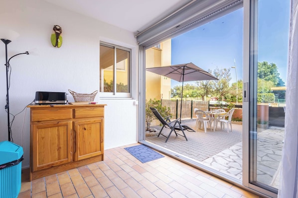 Une pièce avec un meuble en bois, un téléviseur, un panier en osier et une porte-fenêtre coulissante donnant sur une terrasse extérieure avec des chaises et un parasol.