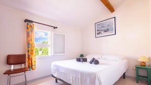 Un dormitorio con cama, una silla, ventana con cortinas, mesita de noche con lámpara y un cuadro en la pared.