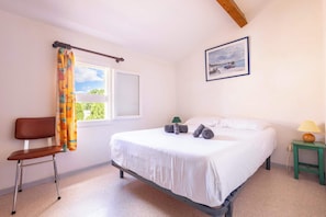 A bedroom with a bed, a chair, a window with curtains, a bedside table with a lamp, and a picture on the wall.
