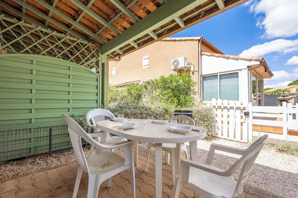 A covered patio with a round table and chairs, a green fence, and a building with a tiled roof in the background.