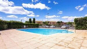 Una piscina con valla metálica, cielo azul despejado y un edificio residencial al fondo.