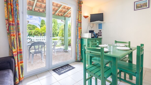 Una habitación con puerta corrediza de vidrio que da a un espacio exterior, mesa de comedor verde con sillas, televisor de pantalla plana en la pared y una pequeña cocina con microondas y cafetera.