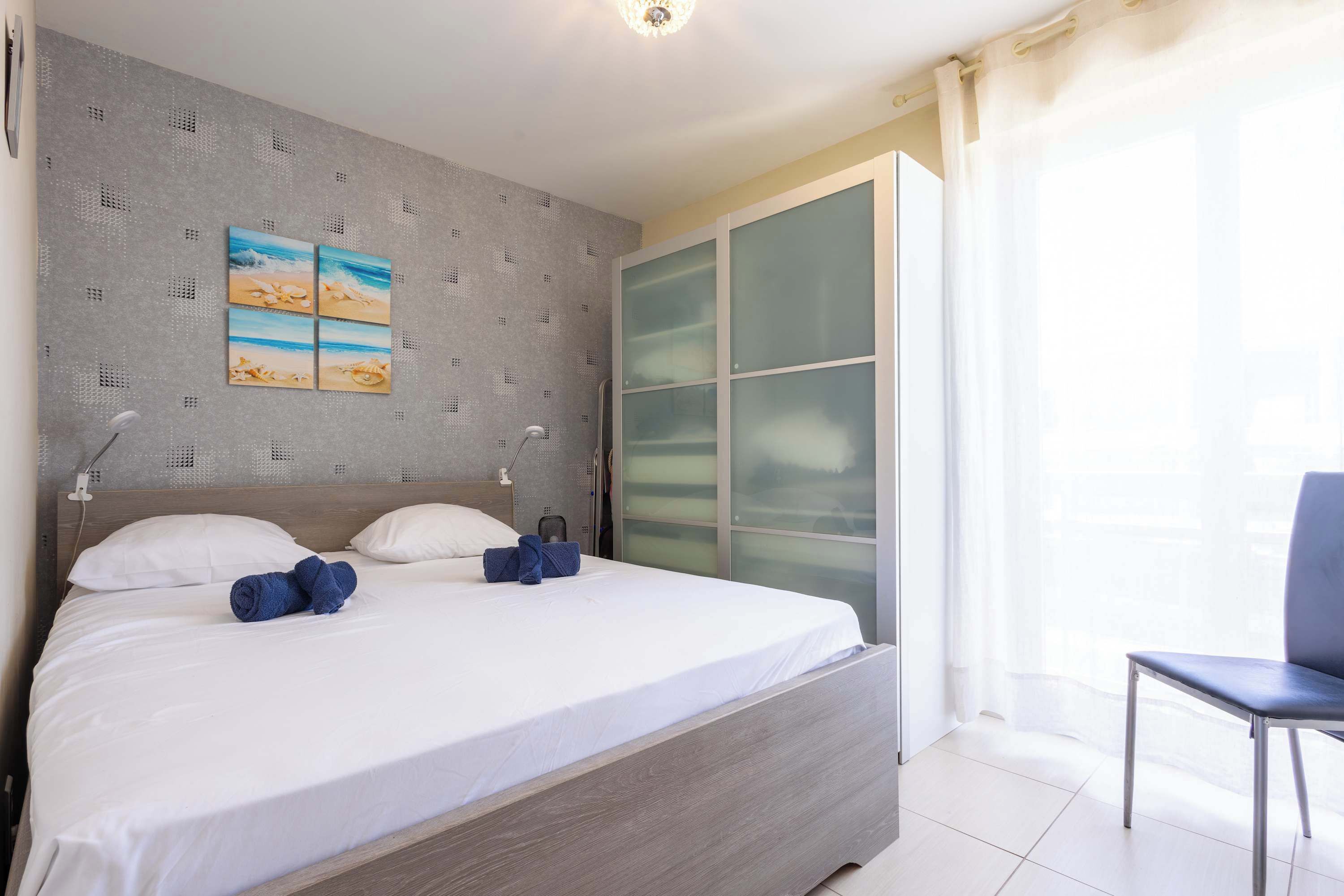Een netjes opgemaakt bed met kussens en handdoeken, een wand met een reliëfpatroon en een schilderij, een glazen schuifdeur en een stoel.