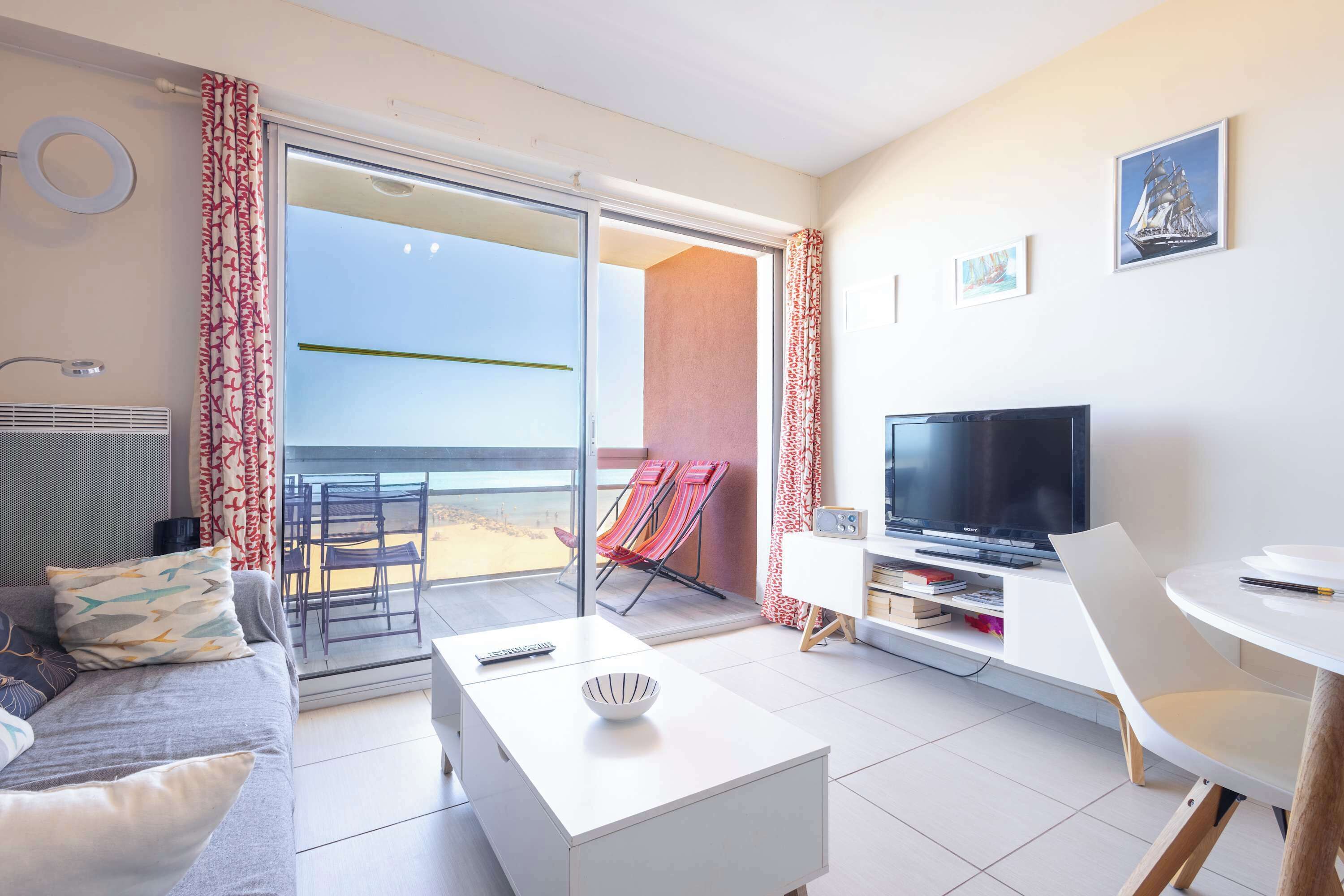 Un salon moderne avec un canapé, une table basse, un téléviseur et un balcon agrémenté de chaises rouges.