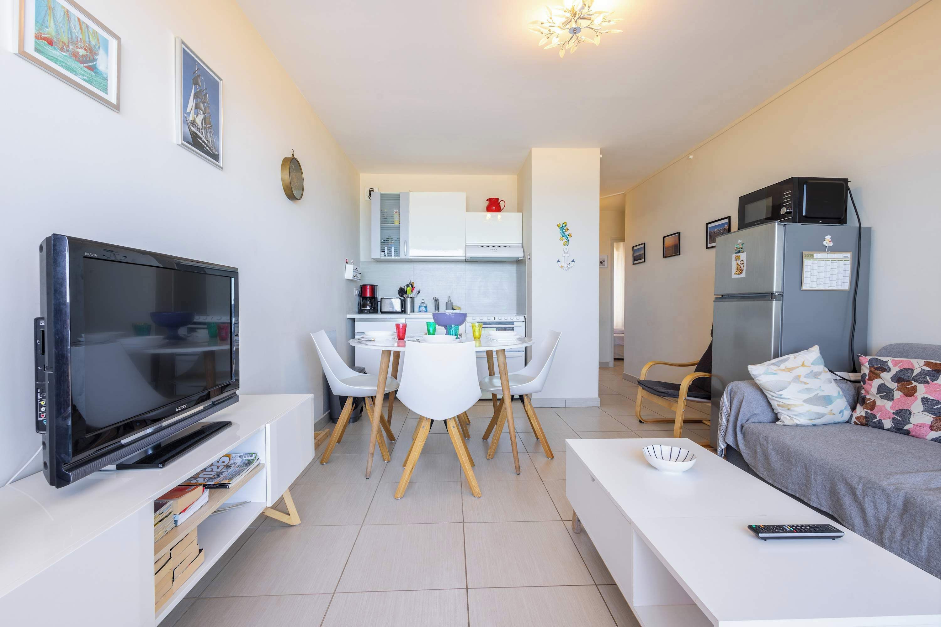 Een moderne woonkamer met een flatscreen-tv, een eethoek met een witte tafel en stoelen, een kitchenette met een koelkast en magnetron, en een bed met geborduurde kussens.