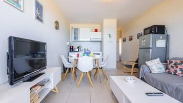 Een moderne woonkamer met een flatscreen-tv, een eethoek met een witte tafel en stoelen, een kitchenette met een koelkast en magnetron, en een bed met geborduurde kussens.