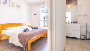 A bedroom with a wooden bed, a bedside table, a nightstand, a window, and a bathroom with a mirror and a towel rack. - Villa Bianca - Air-conditioned pavilion 500m from (Sérignan)