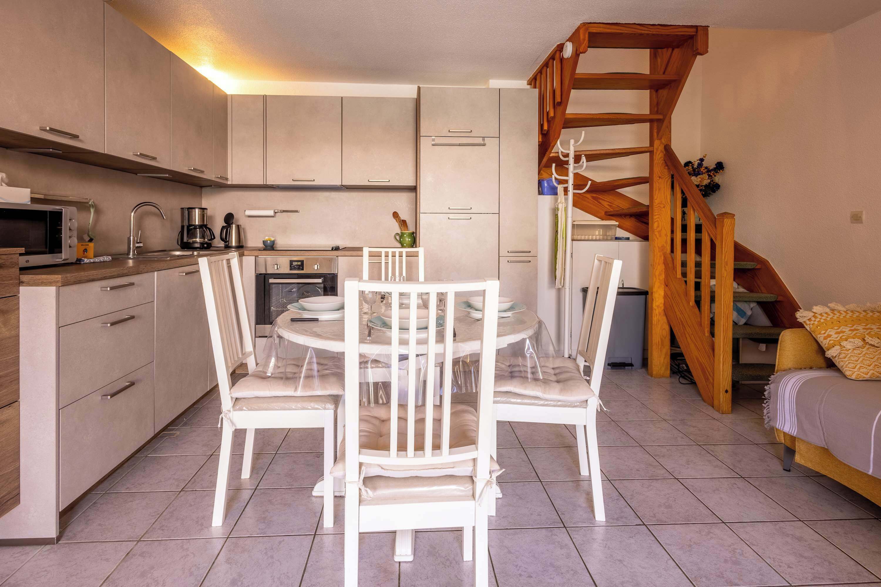 Cocina compacta con electrodomésticos modernos, zona de comedor con sillas blancas y una escalera de madera.