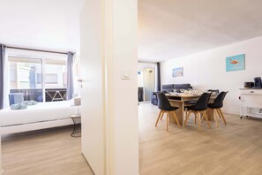 A modern apartment with a bed, a dining area with a table and chairs, a kitchen area, and a view of the city through a large window.