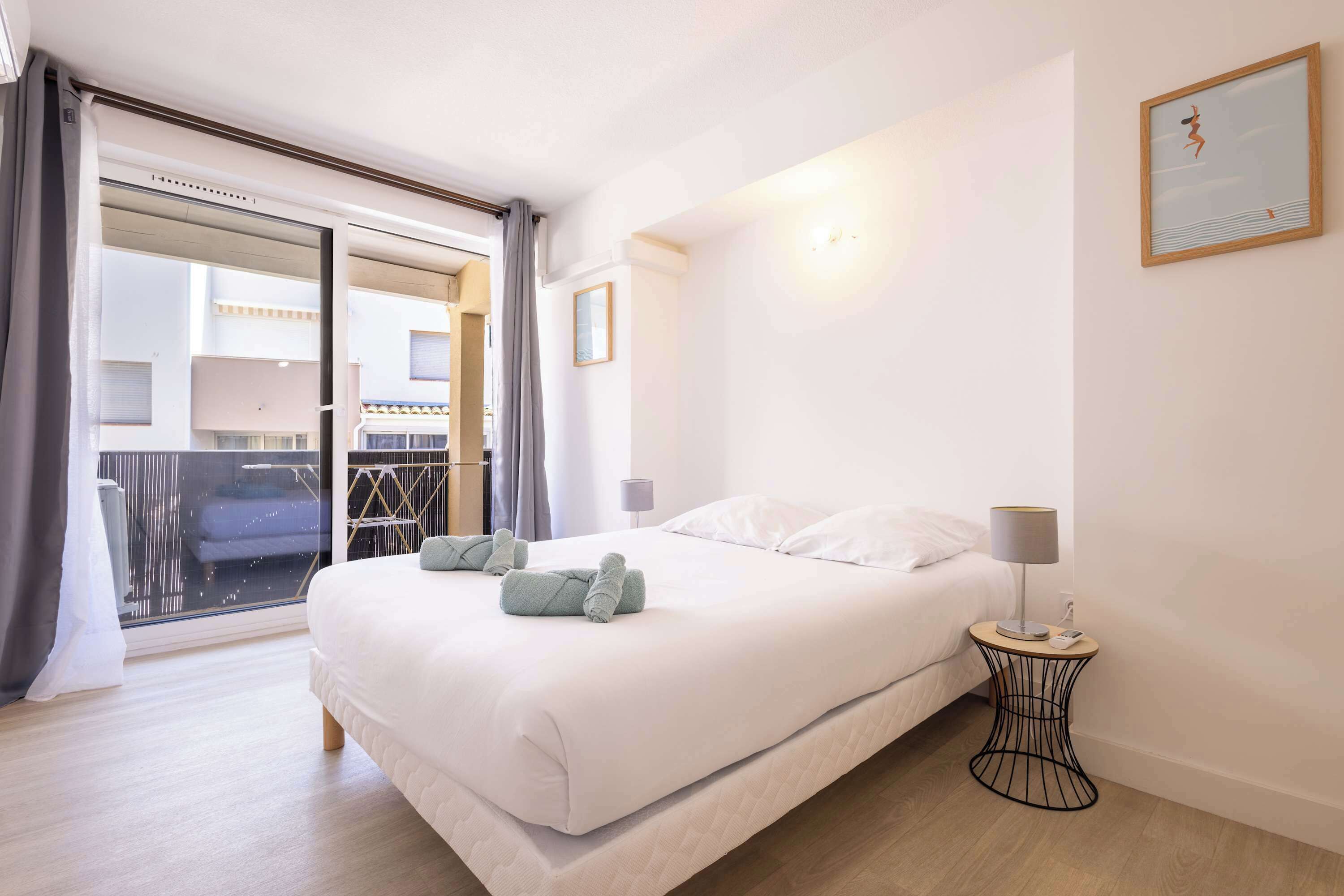 Une chambre d’hôtel avec un lit, une table de chevet, une lampe, un tableau encadré et une vue sur l’extérieur, accessible par une porte coulissante.