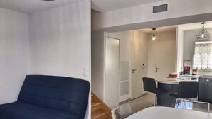 Un salon moderne avec un canapé bleu foncé, une table à manger blanche agrémentée d’un set de table rouge, et un coin cuisine en arrière-plan.