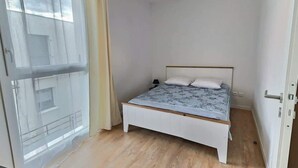Une petite chambre avec un lit simple, un cadre en bois, une couvre-lit de couleur claire, une table de chevet et un luminaire suspendu.