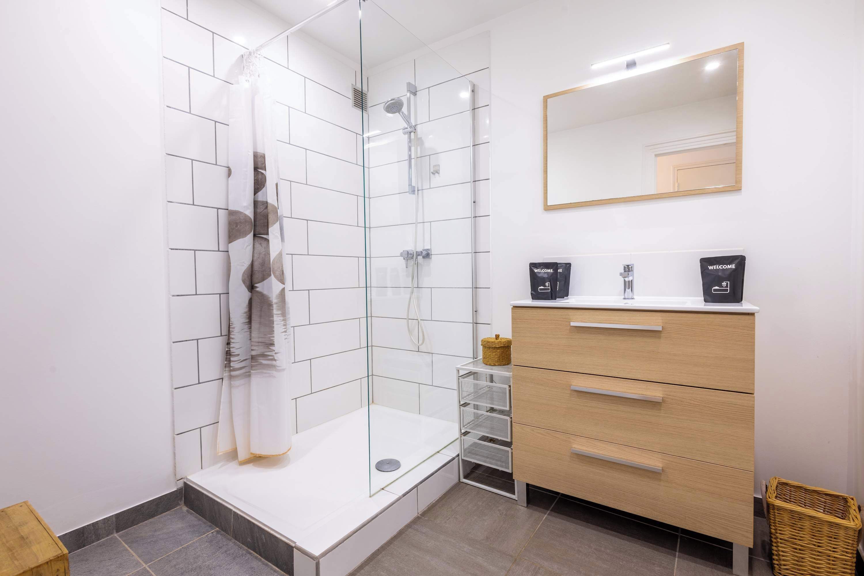 Een moderne badkamer met een douchecabine van glas, een houten wastafelmeubel met een spiegel en een vloer van witte tegels.