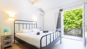 A bedroom with a bed, bedside tables, an air conditioning unit, and a balcony with a view of greenery.