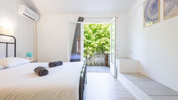 A bedroom with a bed, a nightstand, a lamp, a window, and a view of a balcony with greenery.