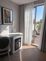 Quarto moderno com escrivaninha, cadeira, forno embutido, janela ampla com cortinas leves e obra de arte emoldurada na parede.