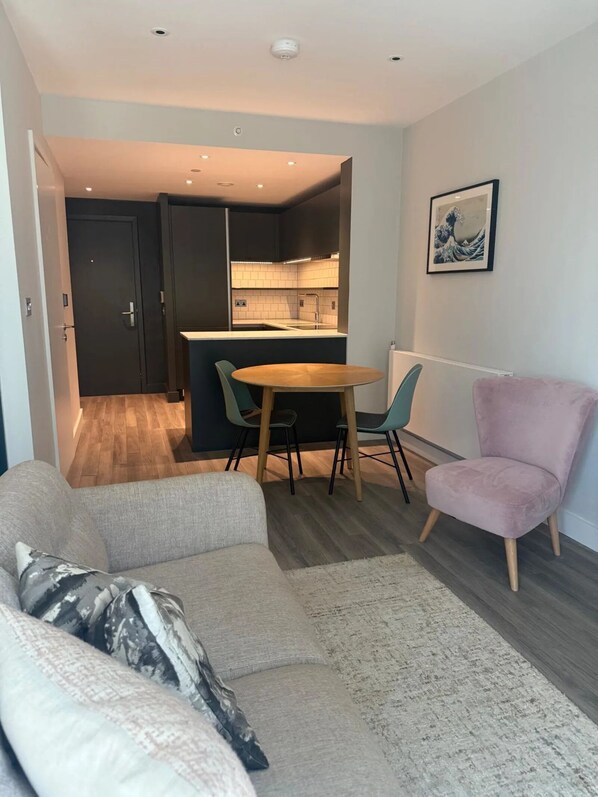 A modern living room with a sofa, a round table, and chairs, a pink chair, and a kitchen visible through an open door. - Alameda - Campus Accommodation (Wembley)