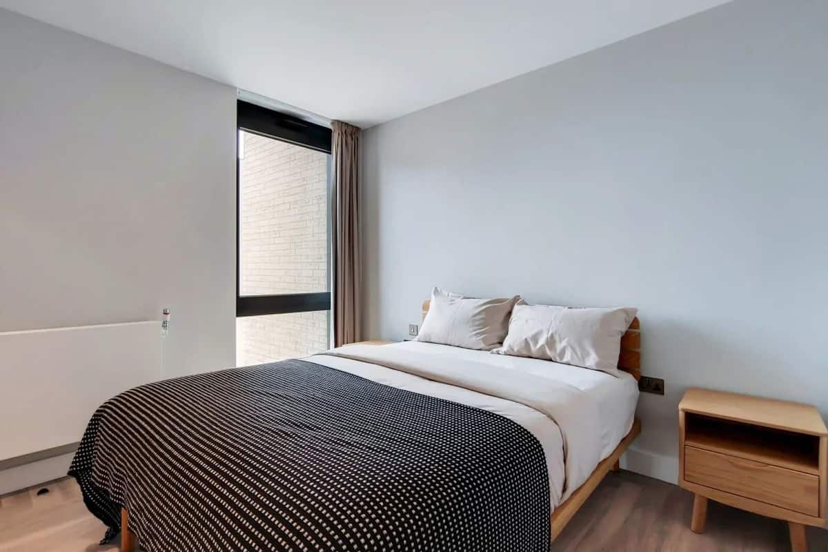A modern bedroom with a large bed, a nightstand, a window with curtains, and a brick wall.