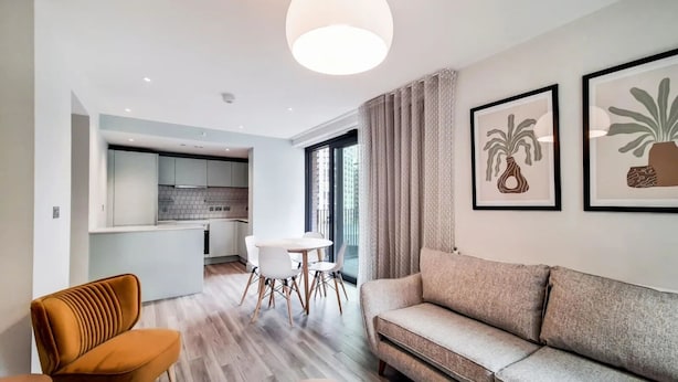 Un salon moderne avec un canapé, une table basse et un coin repas comprenant une table et des chaises. Au fond, on aperçoit une cuisine avec des meubles blancs et une grande fenêtre.