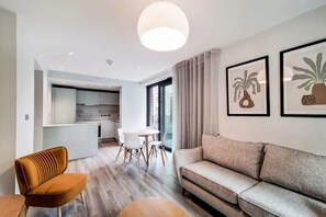A modern living room with a sofa, a coffee table, and a dining area with a table and chairs. There is a kitchen in the background with white cabinets and a large window. - Alameda - Campus Accommodation (Wembley)