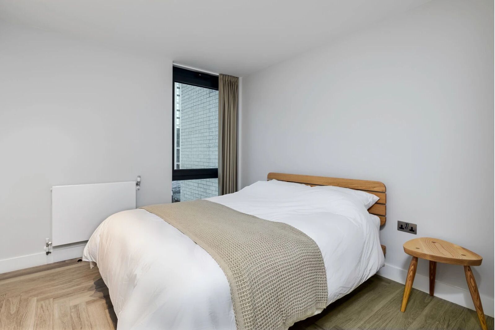 Habitación de hotel moderna con cama de madera, un taburete pequeño de madera, un radiador y una ventana con cortinas.