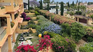 A residential area with a yellow building, a well-maintained garden with various plants and flowers, and a clear sky. - Paradise in Marbella, Sea Sun Golf & more Groudfloor apartment with pool access (Marbella benahavis)