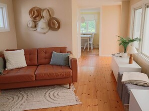 Ein Wohnzimmer mit einem braunen Sofa, einem weißen Teppich, einem Essbereich im Hintergrund und einem Fenster mit einer Pflanze daneben.