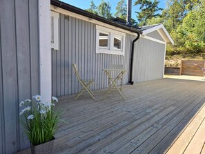A wooden deck with a small table and chairs, a potted plant with white flowers, and a building with a grey exterior and a white window. - 4 Star Holiday Home in Mellbystrand (Mellbystrand)