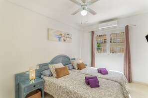 Une chambre avec deux lits, un ventilateur de plafond, un tableau, une fenêtre avec des rideaux et une table de chevet avec une lampe.