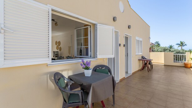 A patio with a table and chairs, a potted plant, and a view of the interior through a window. - Vacation 'Los Mangueros Beach 6 - Mogán' with Mountain View, Wi-Fi and Air Conditioning (Puerto de Mogán)