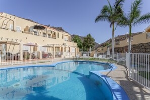 Une piscine à bordures courbes, entourée d’une clôture blanche, et située à côté d’un bâtiment doté de balcons et d’un espace extérieur aménagé pour s’asseoir.