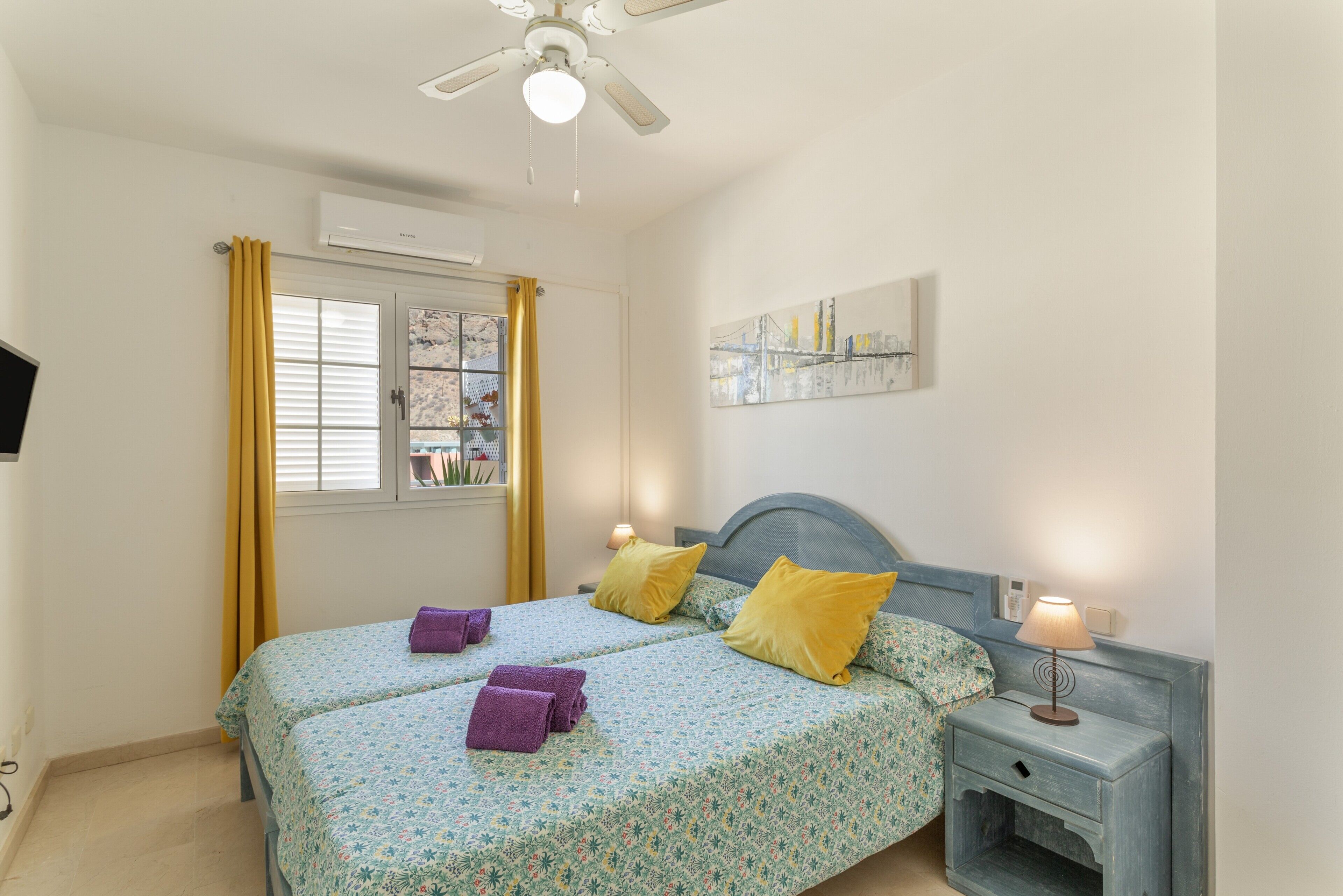 A bedroom with two beds, a ceiling fan, a window with curtains, a painting on the wall, and a bedside table with a lamp.