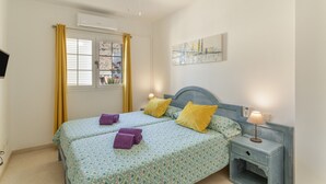 A bedroom with two beds, a ceiling fan, a window with curtains, a painting on the wall, and a bedside table with a lamp.